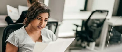 A woman in a gray dress reviews documents at her office desk, with computer screens in the background.
