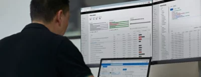 Person vid skrivbordet med tre bildskärmar som visar hotanalys, kontoinloggningar och meddelandeapp.