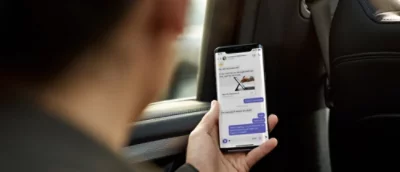 Seseorang dalam mobil melihat smartphone yang menampilkan aplikasi olahpesan dengan beberapa percakapan terlihat.