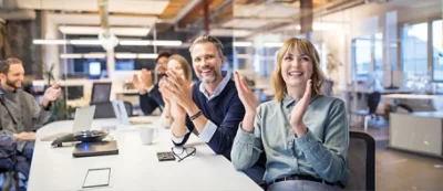 A group of people sit at a conference table in an office, applauding and smiling. Laptops, papers, and a cup are on the table.