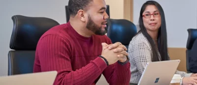 Two people in a meeting: man in red sweater smiles while speaking, woman with glasses listens attentively, both engaged.