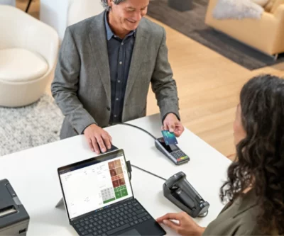 Ein Mann, der an einer Ladenkasse steht und lächelnd auf eine sitzende weibliche Kassiererin blickt, die eine Zahlung mit einem Kartenlesegerät handhabt.