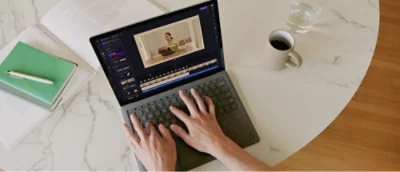 Person using a laptop to edit a video at a round table with a notebook, pen, coffee cup, and glass of water.