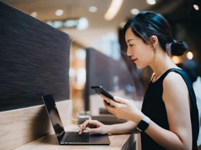 En person sitter vid ett bord med en öppen bärbar dator och en telefon i handen