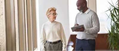 Två personer syns stående inomhus, och en håller i en surfplatta. De verkar vara engagerade i ett samtal. En krukväxt syns i närheten.