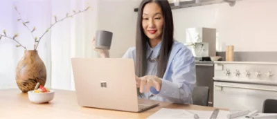 En man med långt hår håller en mugg och använder en bärbar dator vid ett bord. En skål med frukt och en vas syns i närheten.