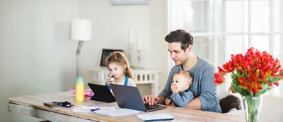 A man works on a laptop at a table with a baby on his lap and a young child beside him using another laptop. Keys, a bottle, and papers are also on the table.