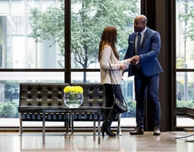Seorang laki-laki dan perempuan berbicara di lobi kantor modern, berdiri di dekat sofa hitam dengan buket bunga kuning