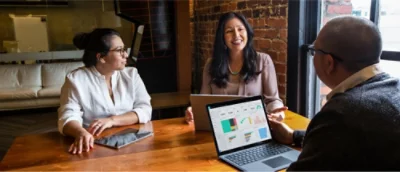 Three people in a business meeting with one person presenting data on a laptop screen.