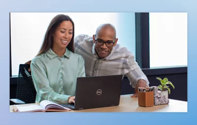 Two individuals collaborating at a desk with a laptop and small potted plants.