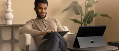 E person i beige tröja sitter i en soffa och håller i en surfplatta. En bärbar dator ligger öppen på ett bord i närheten