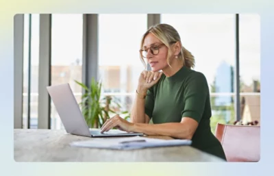 Person working on a laptop at a table near large windows.
