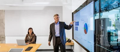 Um homem num fato de trabalho está a apresentar dados num grande ecrã numa sala de conferências, enquanto uma mulher 