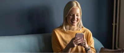 Uma mulher com um suéter amarelo sorrindo enquanto olha para seu smartphone, sentada em um sofá azul.