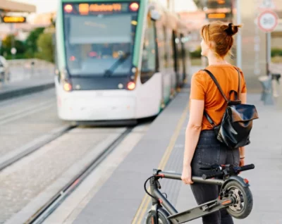 A person with a folded electric scooter and a backpack waits on the sidewalk as a tram approaches.