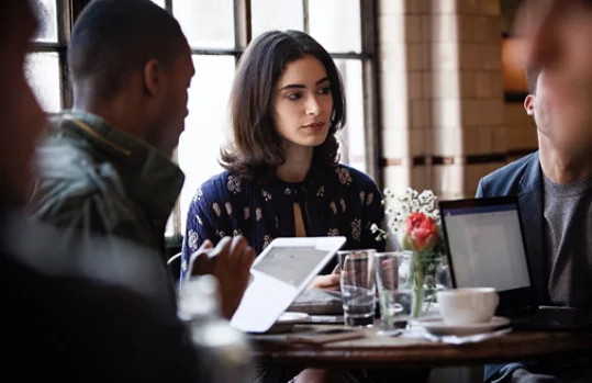 Three people seated at a table in a coffee shop. One person is using a tablet and pen, and another person is using a laptop.