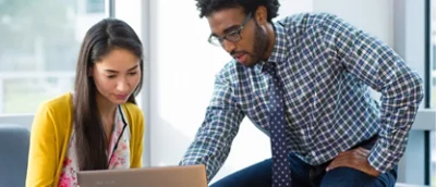 två personer sitter på ett kontor och diskuterar medan de arbetar på en bärbar dator