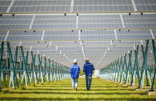 Two people in safety gear walking beneath rows of solar panels mounted on metal structures.
