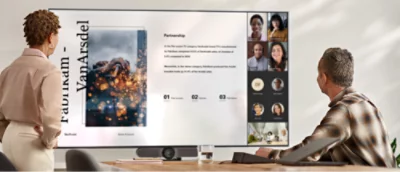 Zwei Personen stehen in einem Besprechungsraum und schauen auf einen großen Bildschirm, auf dem eine virtuelle Besprechung zu sehen ist 