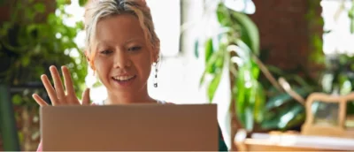 En person sitter i ett ljust rum, ler och vinkar mot en bärbar datorskärm. Växter syns i bakgrunden.