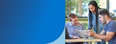 Three individuals engaged in a discussion around a table with a digital tablet, in an outdoor setting.