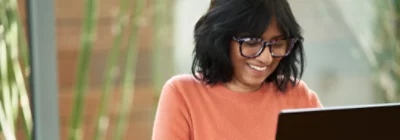 Uma pessoa com óculos e uma camisola cor de laranja sorri enquanto olha para um portátil. 