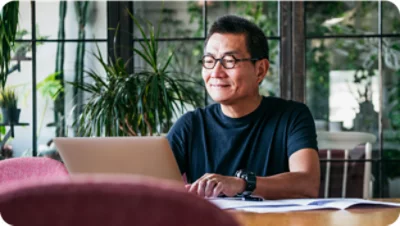 A person wearing glasses and a black shirt sits at a desk, smiling while looking at a laptop. 
