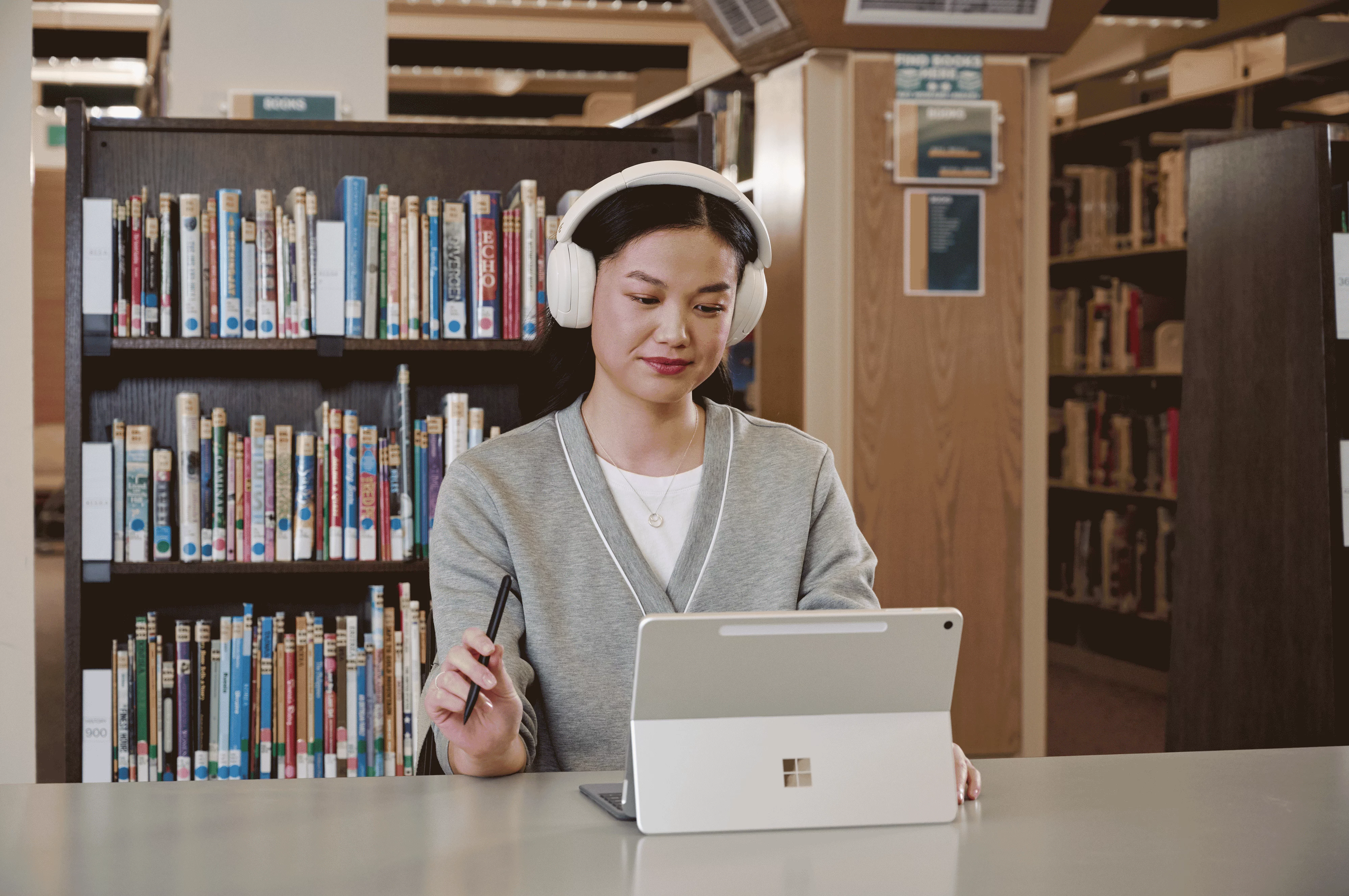 A person wearing large headphones and using a Surface device with a pen while seated at a table in a library