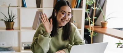 En person i en grön tröja och glasögon sitter vid ett skrivbord framför en bärbar dator och ler och vinkar.