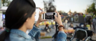 A person takes a photo with a smartphone at an outdoor event, with people and colorful decorations visible in the background.