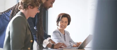 Tre personer i ett mötesrum, varav en sitter och tittar på en bärbar dator medan de två övriga står i närheten